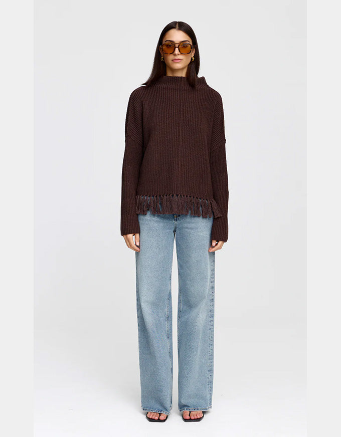 A model wearing a dark brown ribbed knit sweater with fringe trim, paired with light-wash wide-leg jeans and sunglasses, standing against a light gray studio background.