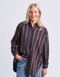 A smiling blonde woman wearing a brown button-up shirt with vertical blue and red stripes, paired with blue jeans, against a light gray studio background.