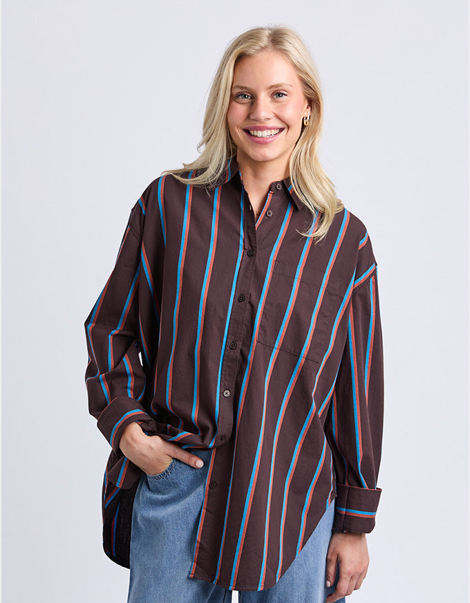 A smiling blonde woman wearing a brown button-up shirt with vertical blue and red stripes, paired with blue jeans, against a light gray studio background.