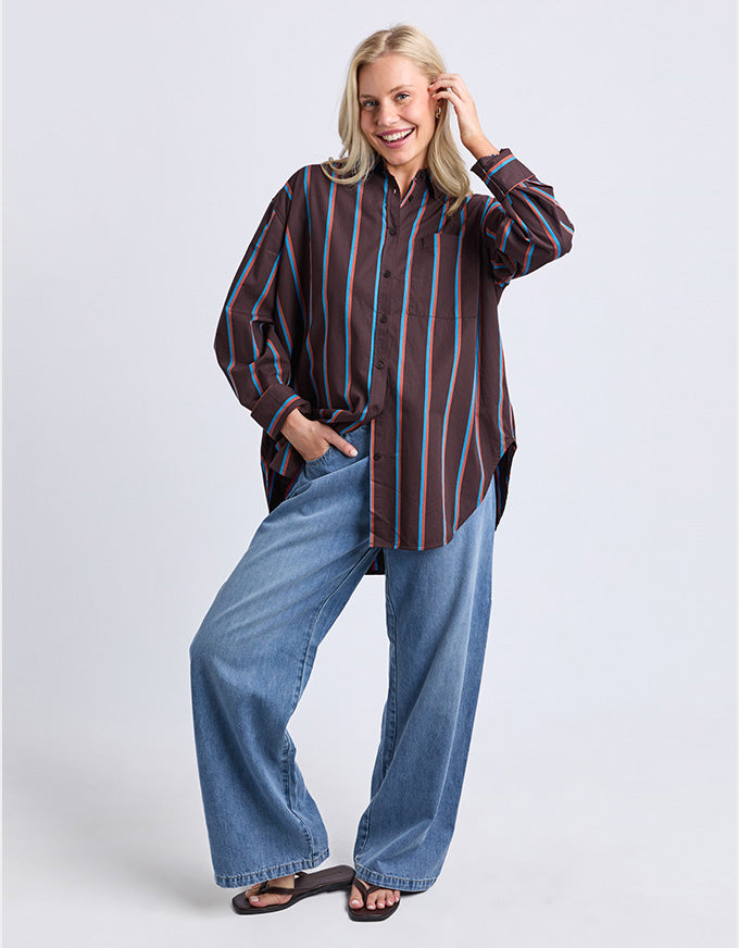 A woman modeling an oversized long-sleeve button-up shirt with vertical stripes in dark brown, black, and light blue, paired with wide-leg blue jeans and black sandals, against a light gray studio background.