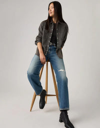 Woman wearing a gray shirt, black top, blue jeans, and black boots on a white background