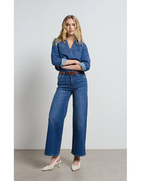 Woman wearing a denim shirt and jeans standing against a plain background