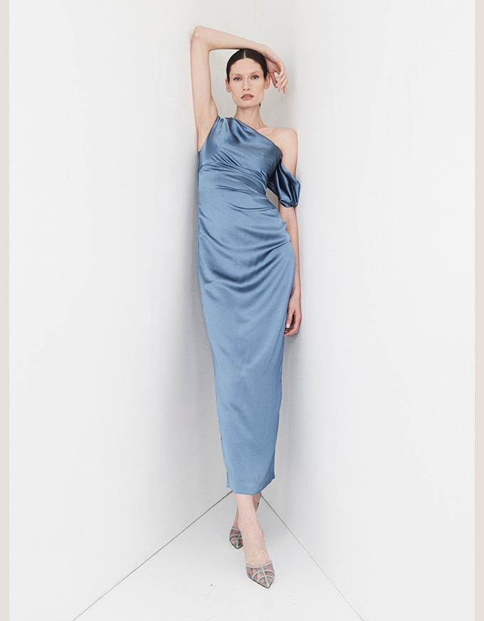 A model wearing a light blue satin one-shoulder midi dress with a draped bodice and skirt, posing in a minimalist white studio corner.