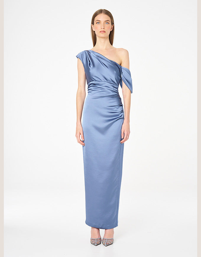 A woman modeling a floor-length, one-shoulder satin dress in dusty blue, paired with patterned high heels, against a plain white studio background.