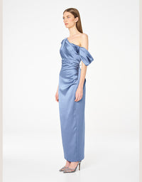 A woman modeling a floor-length, one-shoulder satin gown in dusty blue. The dress features a dramatic gathered puff sleeve and a ruched bodice.