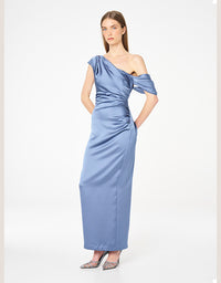 A woman modeling a floor-length, one-shoulder satin dress in dusty blue, styled with metallic pointed-toe heels against a plain white studio background.