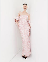 A model wearing a strapless light pink off-the-shoulder gown with a textured floral jacquard pattern, standing in a minimalist white studio corner.