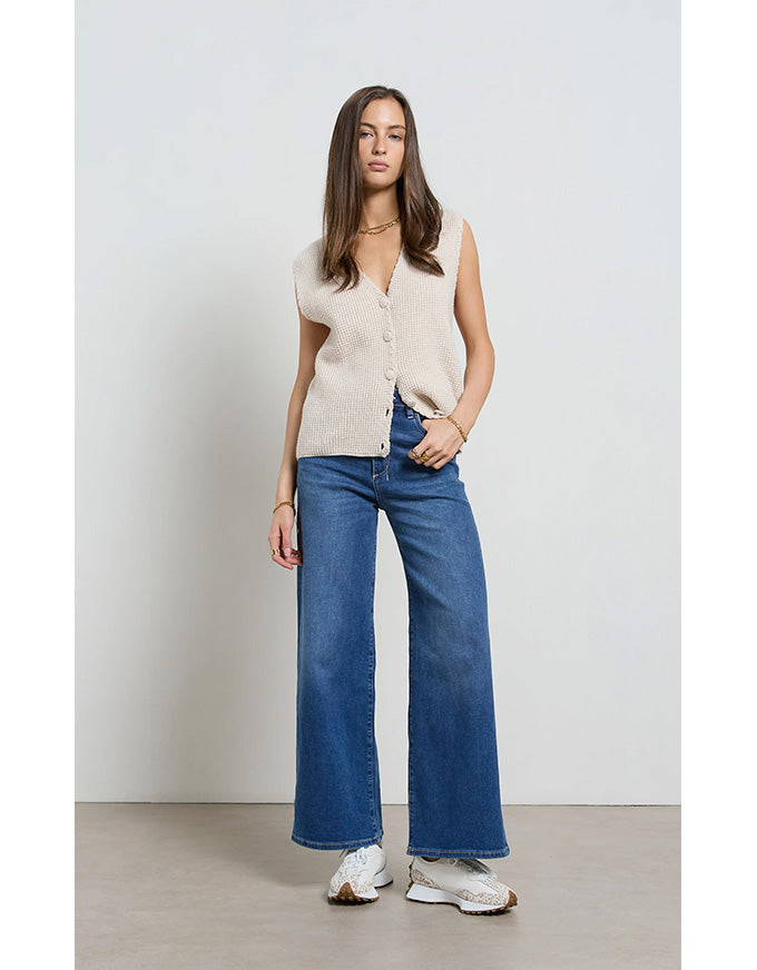 Woman wearing a sleeveless beige top and blue jeans against a white background