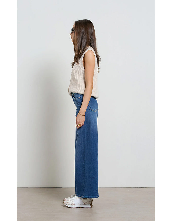 Woman wearing a beige sleeveless top and blue jeans standing against a white wall.