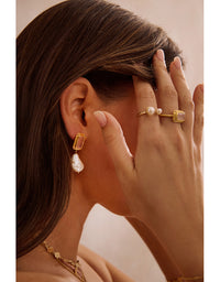 Close-up of a woman wearing gold earrings and a ring with a pearl, against a neutral background.