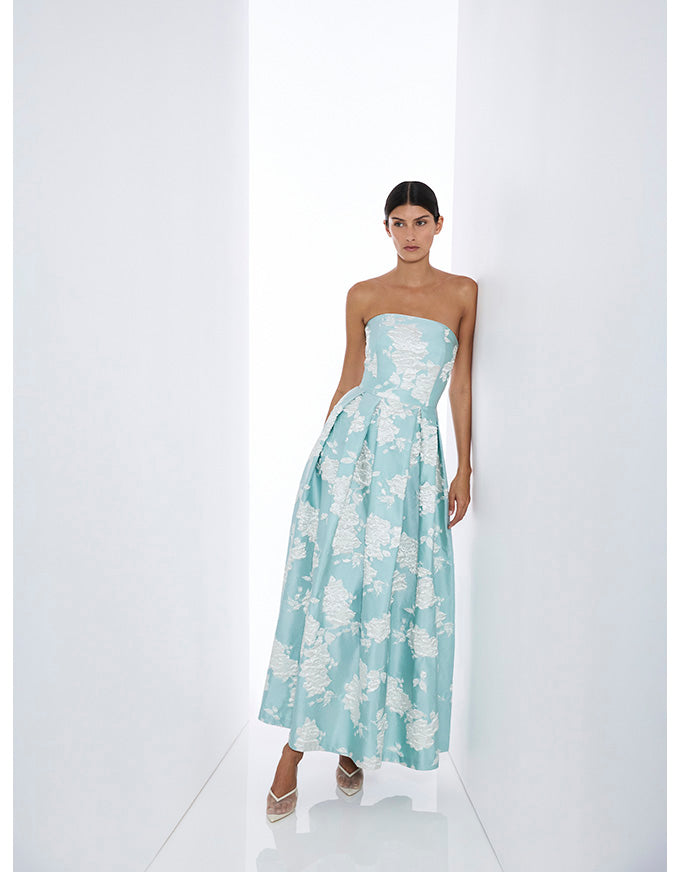 A woman modeling a strapless light blue maxi dress with a white floral jacquard pattern, standing in a minimalist white studio setting.