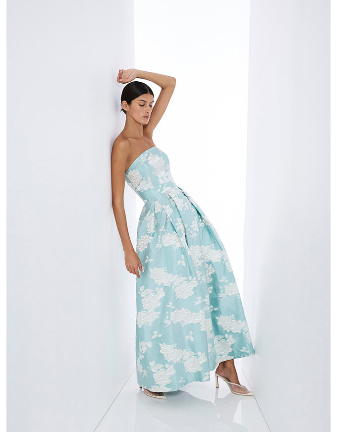A woman models a strapless light blue maxi dress with a white floral pattern, standing in a bright white studio setting.