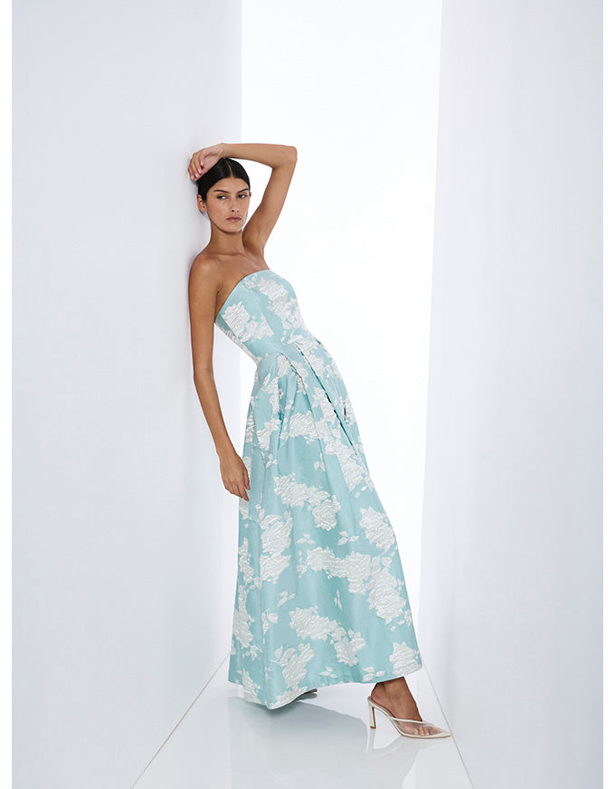 A woman models a strapless light blue maxi dress with a white floral pattern, leaning against a white wall in a minimalist white room.