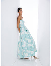 A woman models a strapless floor-length gown with a light aqua blue and white floral jacquard pattern, standing in a minimalist white studio setting.