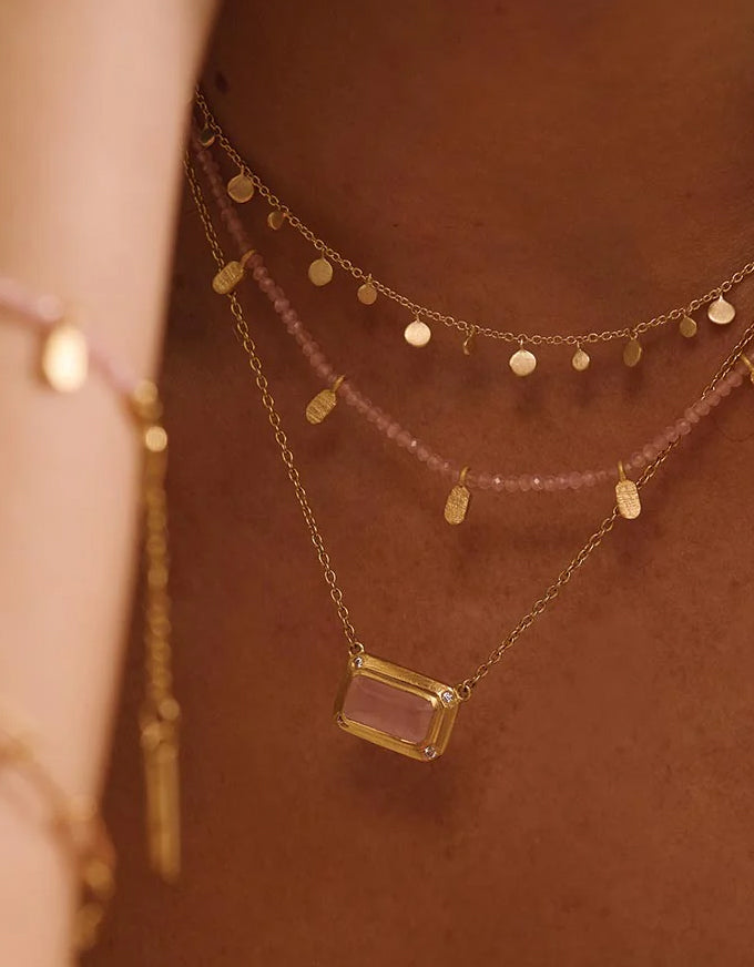 Gold necklace with a rectangular pendant on a person's neck against a brown background