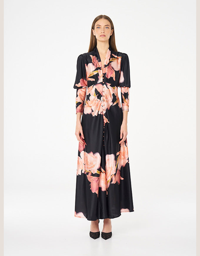 A woman modeling a long-sleeved black maxi dress with a pink and peach floral print, standing against a plain white studio background.