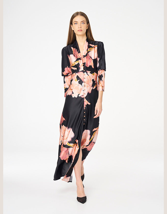 A woman modeling a black satin-like midi dress with a pink and peach floral print, standing against a plain white studio background.