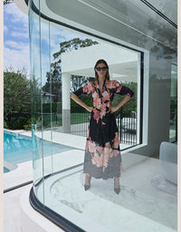 A woman wearing a black midi dress with a pink and peach floral print stands with hands on hips inside a modern glass-walled structure overlooking a swimming pool.