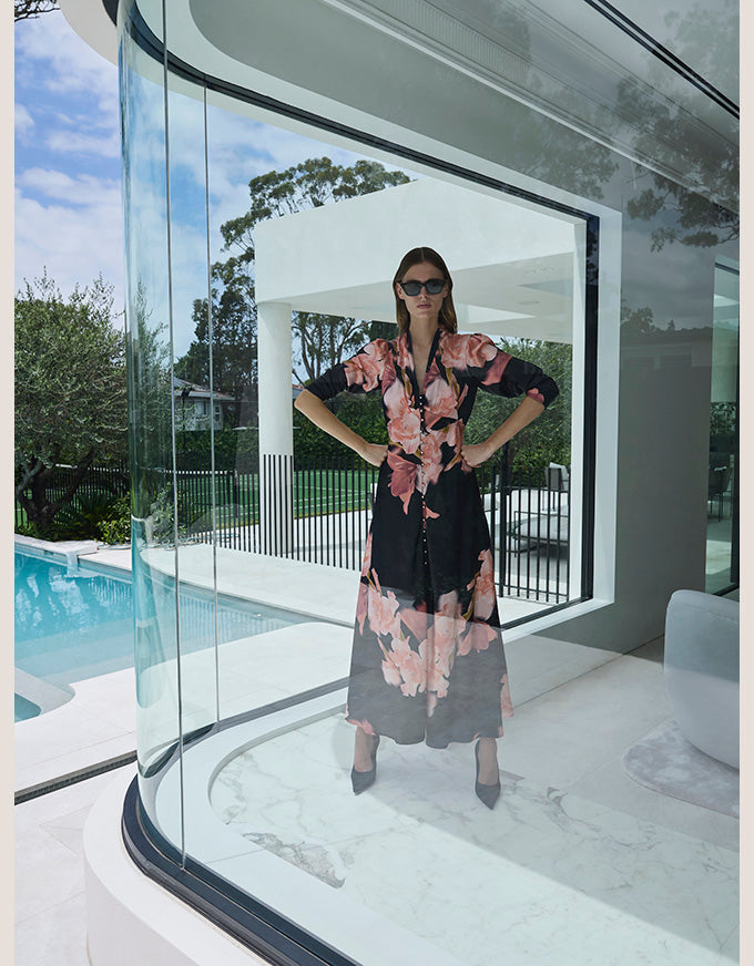 A woman wearing a black midi dress with a pink and peach floral print stands with hands on hips inside a modern glass-walled structure overlooking a swimming pool.