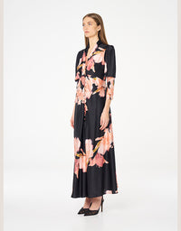 A woman modeling a black midi dress with a pink and peach floral print, featuring short sleeves, a button-up front, and a V-neckline, set against a plain white studio background.