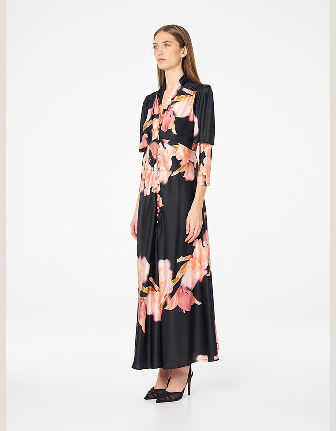 A woman modeling a black midi dress with a pink and peach floral print, featuring short sleeves, a button-up front, and a V-neckline, set against a plain white studio background.