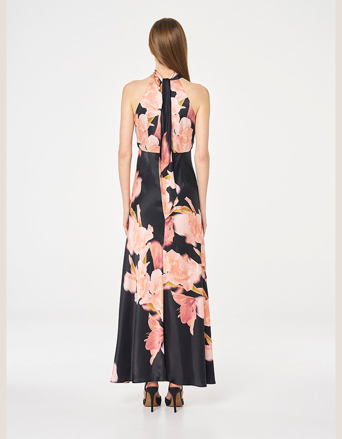 A woman viewed from the back wearing a sleeveless halter-neck maxi dress with a black base and large pink floral print, paired with brown heels, against a plain light gray studio background.