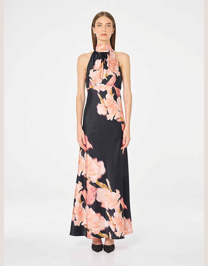 A model wearing a sleeveless halter-neck maxi dress with a black base and a large pink floral print, standing against a plain white studio background.