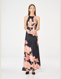 A model wears a sleeveless halter-neck maxi dress featuring a black base with a large pink floral print, set against a plain light gray studio background.