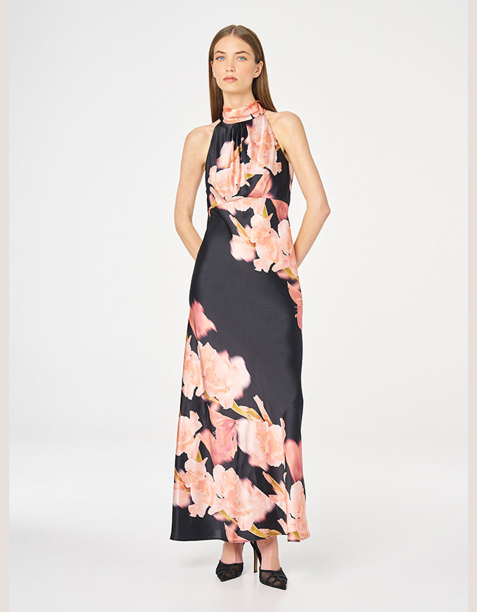 A woman modeling a sleeveless halter-neck maxi dress with a black base and a large pink floral print, standing against a light gray studio background.