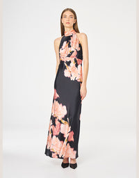 A woman modeling a sleeveless halter-neck maxi dress with a black base and a large pink floral print, set against a plain light gray studio background.
