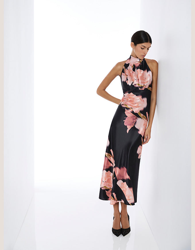 A woman models a sleeveless black midi dress with a pink floral print, standing in a minimalist white studio setting.