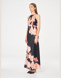 A woman modeling a sleeveless high-neck maxi dress with a black base and a large pink floral print, photographed against a plain light gray studio background.