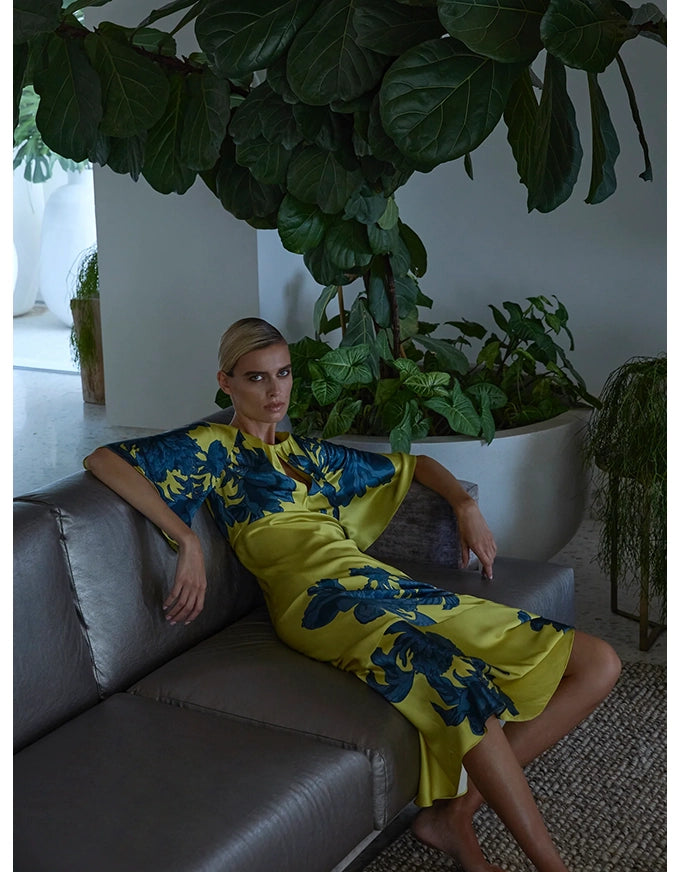 Woman in a yellow and blue dress sitting on a couch with plants in the background