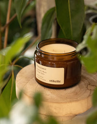 Candle in a brown jar with a label on a wooden surface surrounded by green leaves