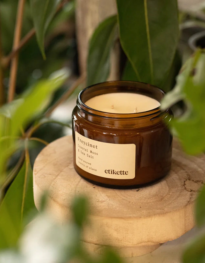 Candle in a brown jar with a label on a wooden surface surrounded by green leaves