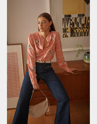 Woman in a pink blouse and blue jeans sitting on a wooden surface with a minimal background