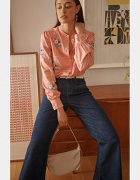 Woman wearing a pink embroidered blouse and blue jeans sitting on a wooden bench.