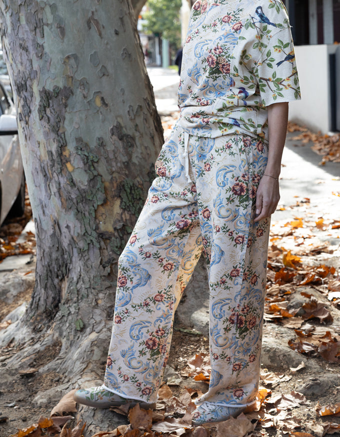 A person wearing a matching short-sleeve top and wide-leg trousers pajama set with a vintage floral and bird print, standing outdoors next to a large tree trunk on a leaf-covered ground.