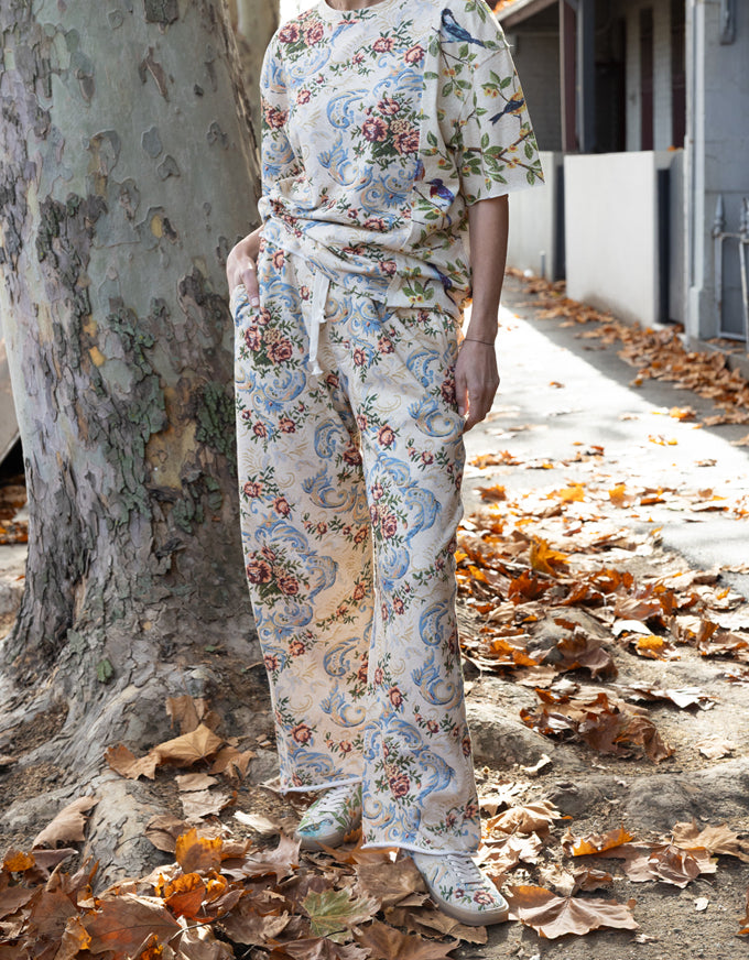 A person wearing a matching short-sleeve tie-front top and wide-leg pants set with a blue, green, and pink floral paisley print, standing outdoors next to a large tree with fallen leaves on the ground.