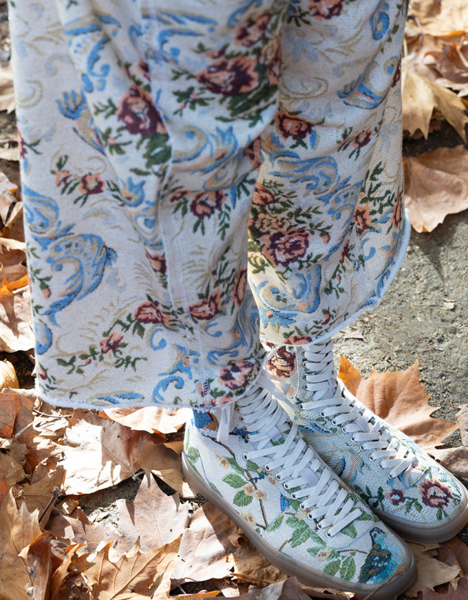 A close-up of a person's legs and feet wearing white high-top sneakers and matching white pants with a colorful floral and bird pattern, standing outdoors on dry brown autumn leaves.