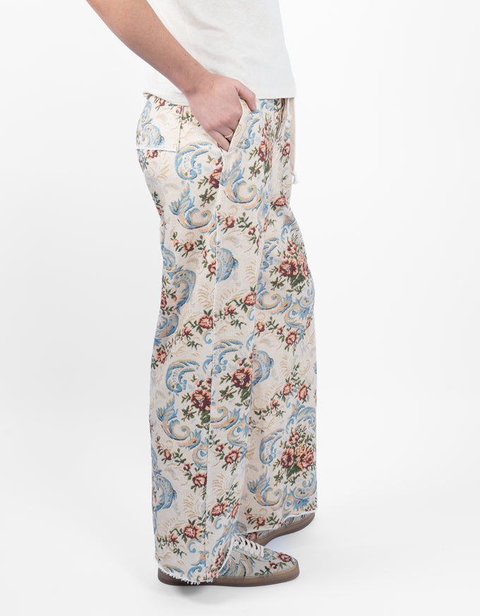 A person wearing wide-leg trousers with a detailed floral and scroll pattern in blue, red, and green on a cream background, paired with a white top and brown shoes, shown against a plain white studio background.