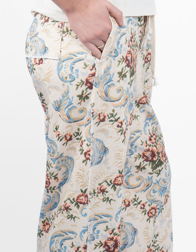 Close-up of a person wearing patterned trousers, showing a hand pulling the zipper and the fabric's ornate floral and scroll design in blue, beige, and brown tones.