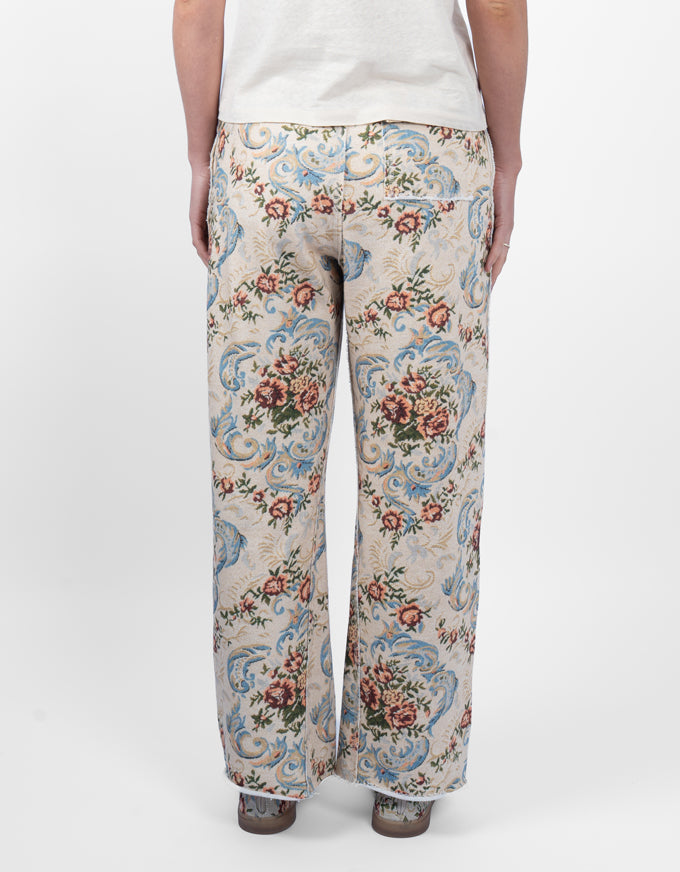 A person shown from the back wearing wide-leg trousers with an ornate floral and scroll pattern in beige, blue, pink, and green, paired with a white top and brown shoes, against a plain white studio background.