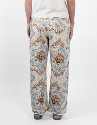 A person shown from the back wearing wide-leg trousers with an ornate floral and scroll pattern in beige, blue, pink, and green, paired with a white top and brown shoes, against a plain white studio background.