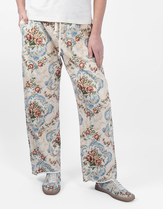 A person wearing wide-leg trousers with an ornate floral and scroll pattern in cream, blue, pink, and green, paired with matching patterned sneakers against a plain light background.