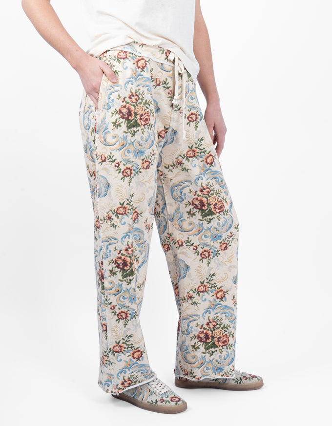 A person wearing loose, wide-leg pajama pants with a vintage-style floral and scroll pattern in cream, light blue, pink, and green, paired with a white top and gray slip-on shoes against a plain white background.