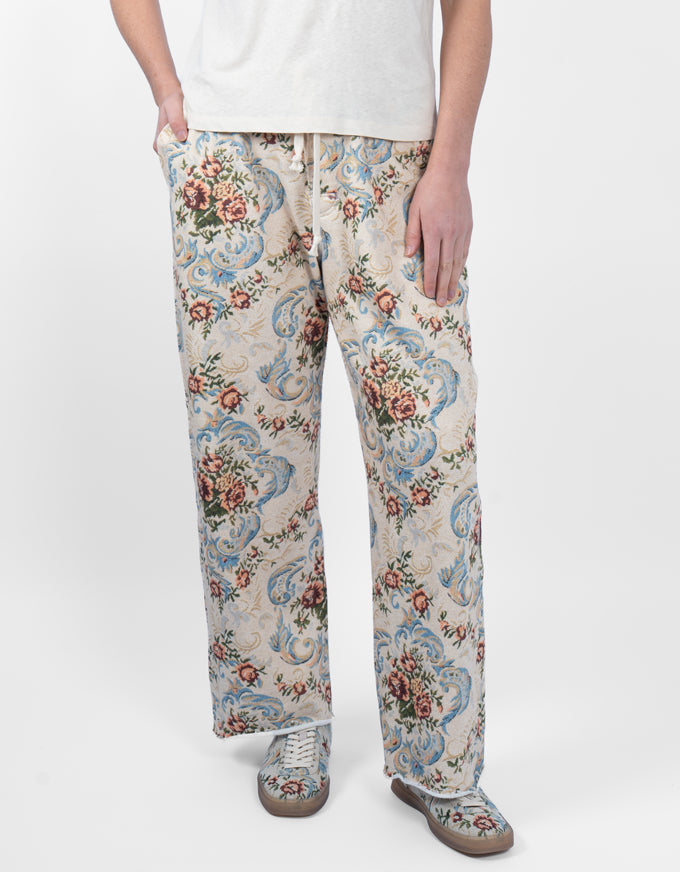 A person wearing loose, wide-leg pants with an ornate floral and scroll pattern in beige, blue, pink, and green, paired with a white top and gray slip-on shoes, photographed against a light gray studio background.