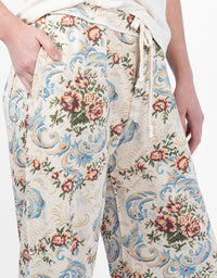 Close-up of a person wearing wide-leg trousers with a cream base and an ornate floral and scroll pattern in blue, pink, green, and tan. The pants have a drawstring waist and side pockets, and the model's hands are visible.