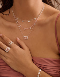 Close-up of a person wearing multiple necklaces, rings, and bracelets on a neutral background.