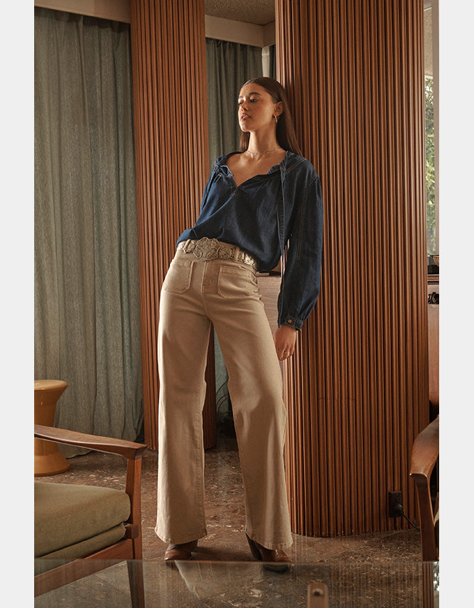 Woman wearing a blue shirt and beige pants standing in a room with wooden walls and furniture.